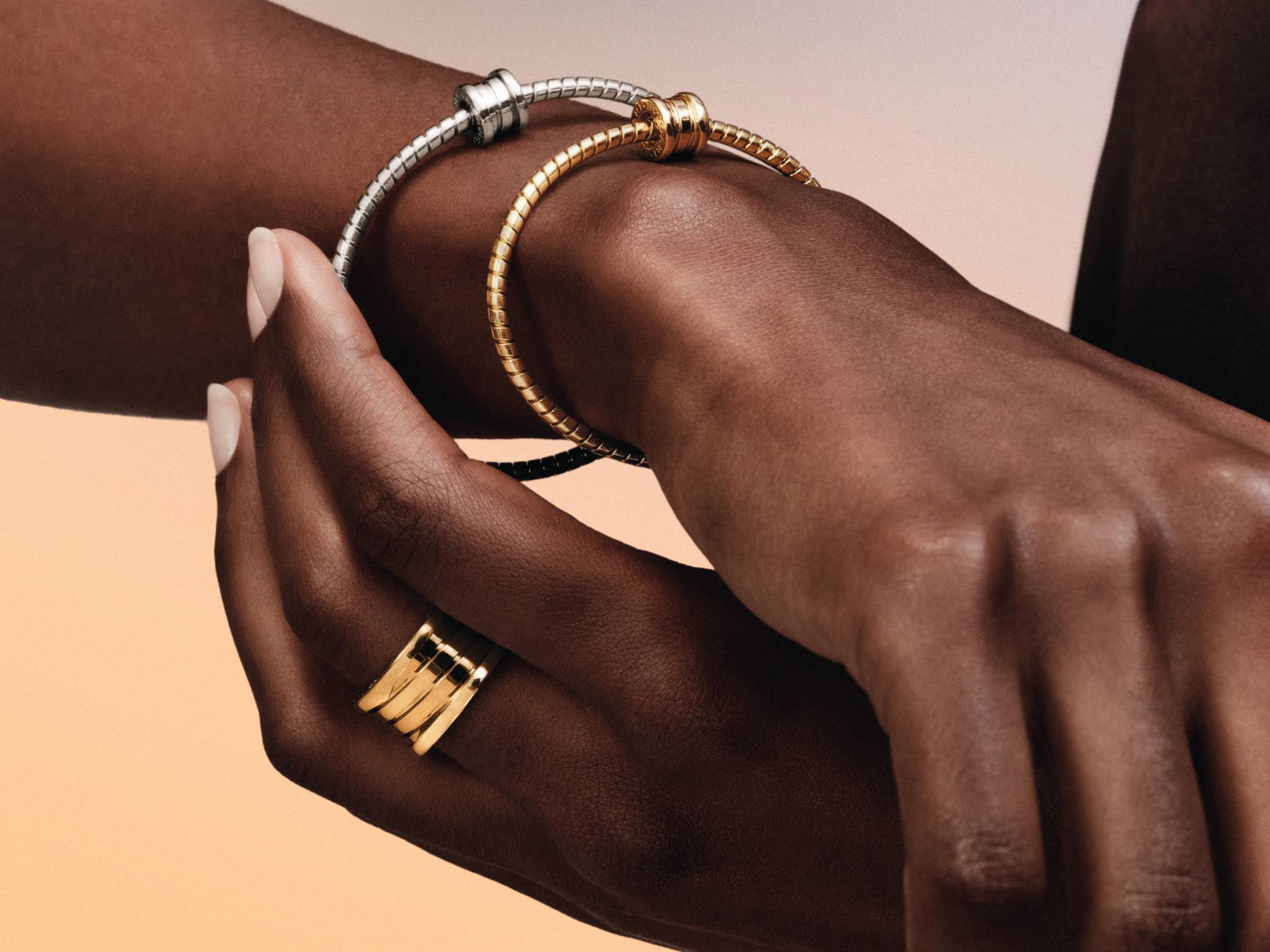 Deux mains ornées de bracelets jonc en or et argent et d’une bague dorée multi-anneaux, sur fond beige.