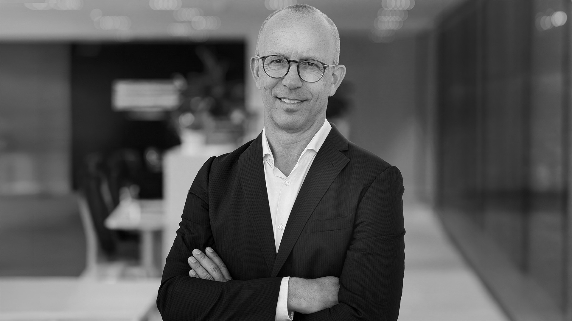 Homme chauve souriant en veste noire et chemise blanche, bras croisés, portrait en noir et blanc dans un bureau moderne.