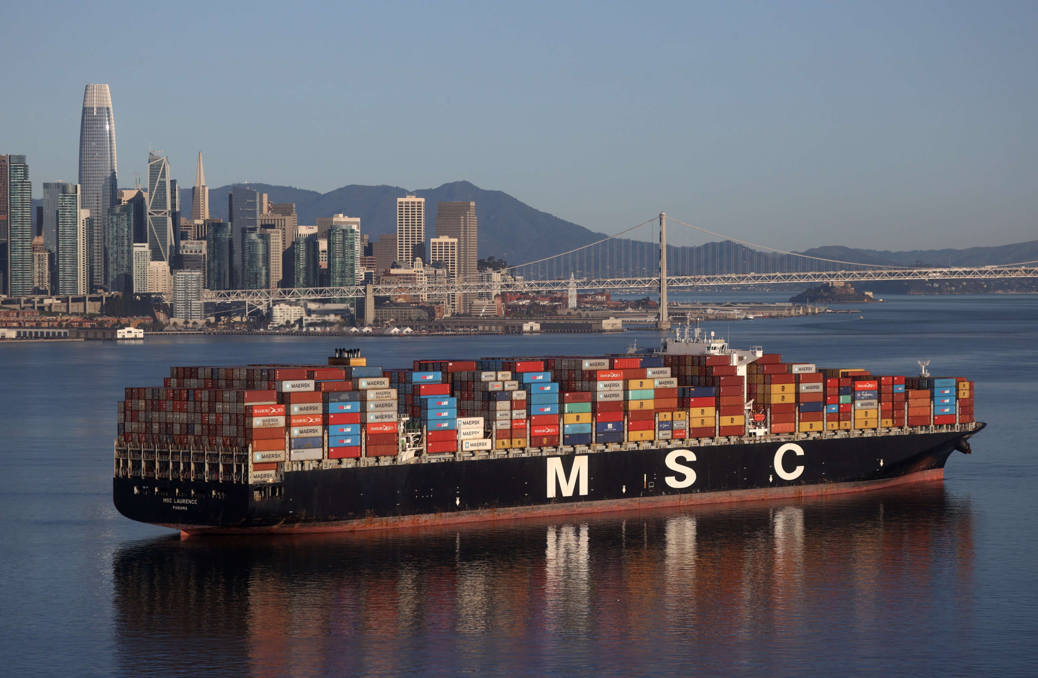 Un porte-conteneurs MSC chargé à pleine capacité dans la baie de San Francisco, avec le port d’Oakland et le skyline de San Francisco en arrière-plan.