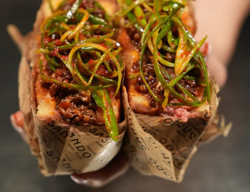 Deux sandwichs garnis de viande épicée, sauce et oignons verts, enveloppés dans du papier brun imprimé, tenus dans les mains.