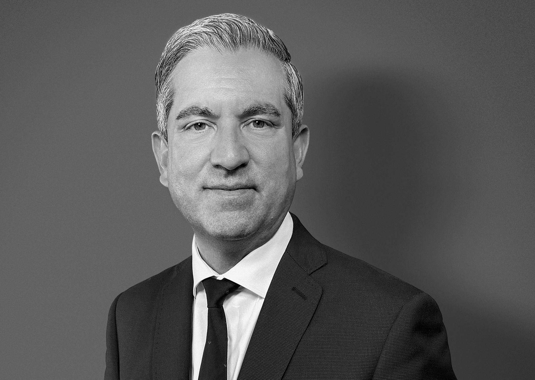 Portrait en noir et blanc d’un homme en costume sombre et cravate, aux cheveux courts grisonnants, sur fond gris neutre.