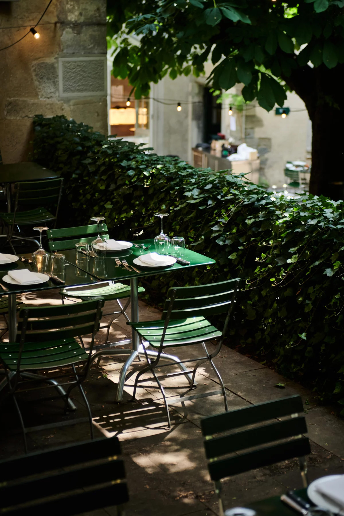 Terrasse de restaurant avec tables et chaises vertes dressées, entourée de feuillage luxuriant et guirlandes lumineuses.