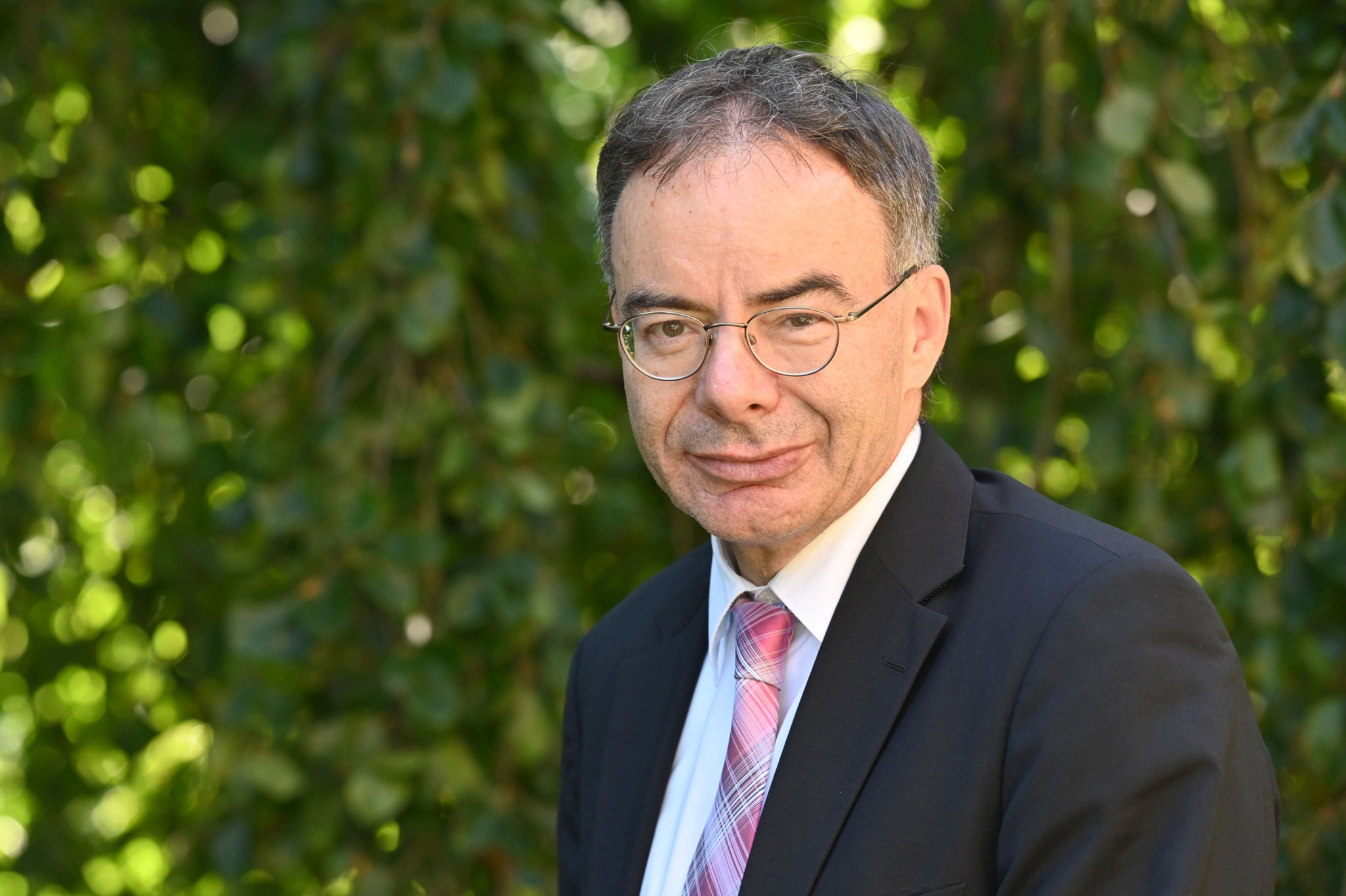 Homme en costume sombre et cravate rose, portant des lunettes, devant un fond de feuillage vert.