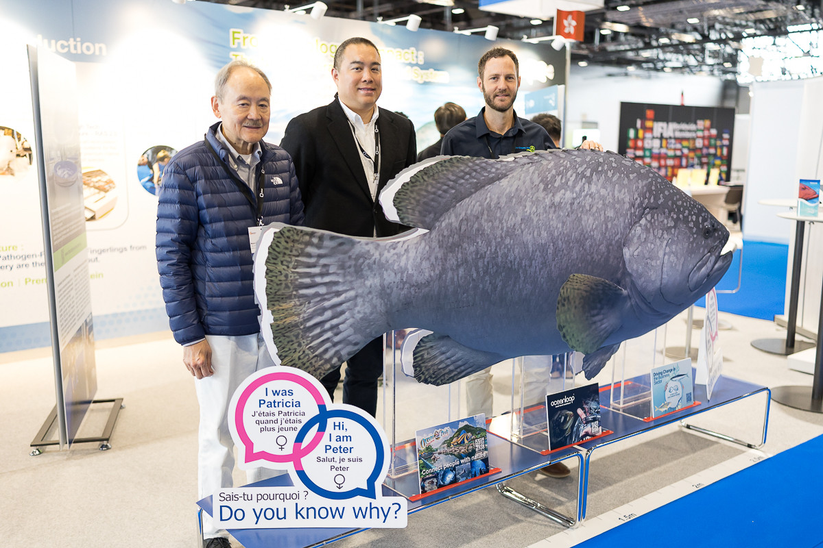 Trois hommes posent à côté d’une grande maquette de poisson bleu-gris dans un salon professionnel, avec un panneau bilingue ’Do you know why?’