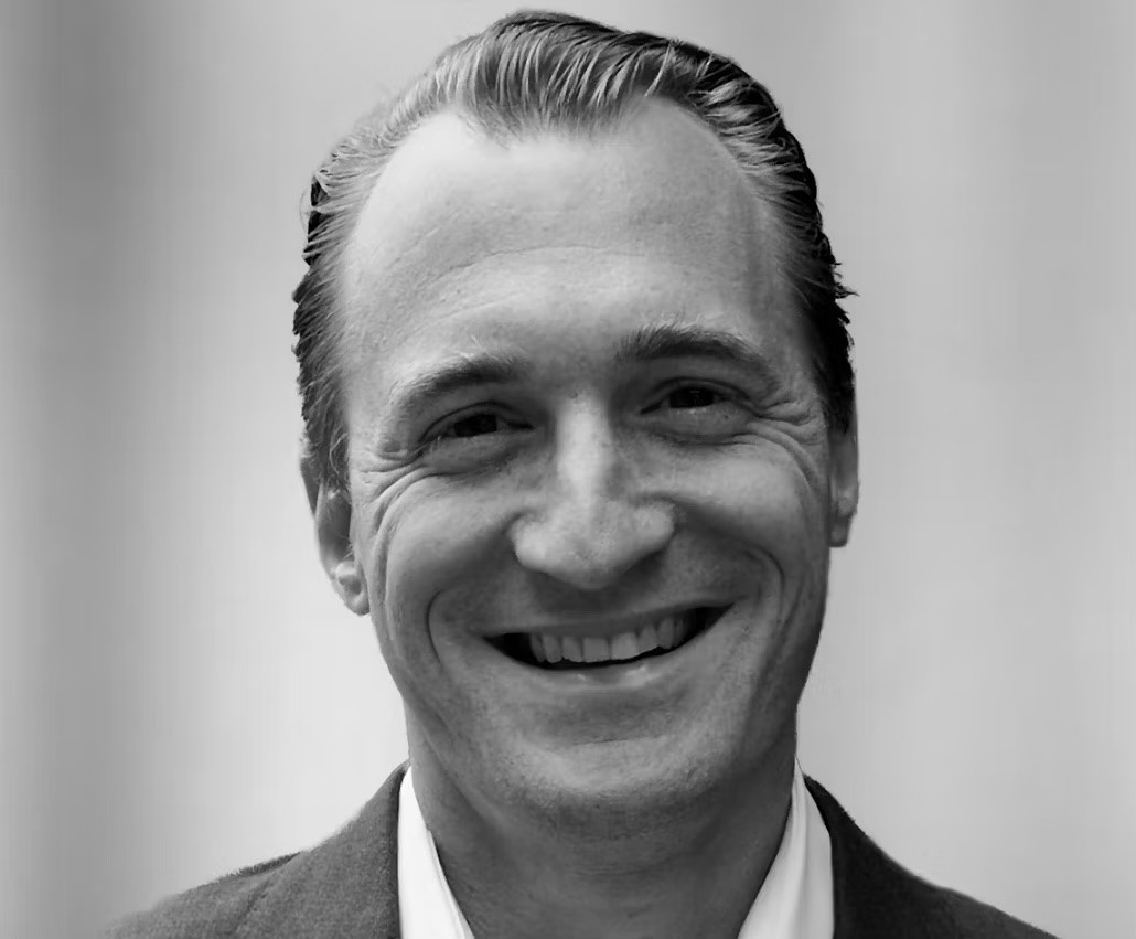 Portrait en noir et blanc d’un homme souriant, cheveux courts coiffés en arrière, portant un veston sombre.