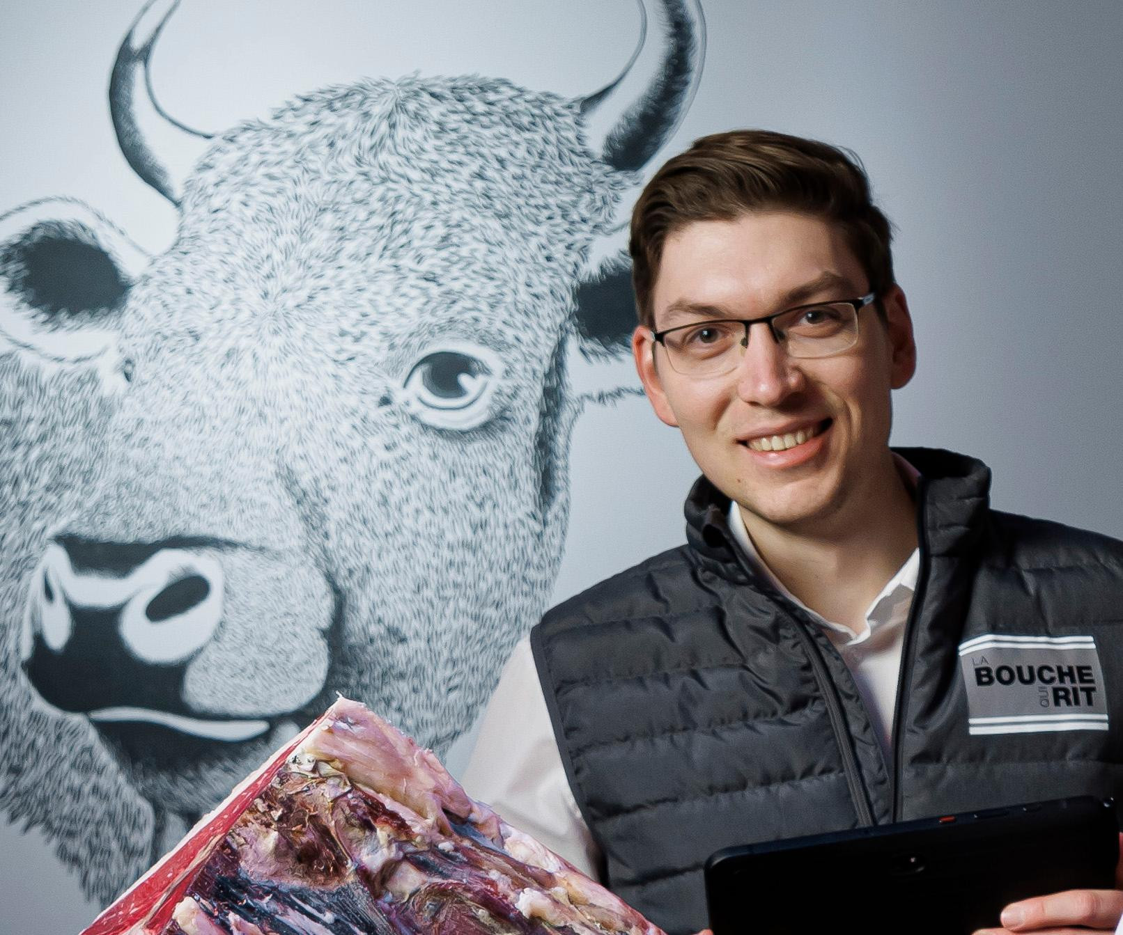 Un boucher souriant portant un gilet ’Boucherit’ tient une pièce de viande devant une fresque murale représentant un bison.