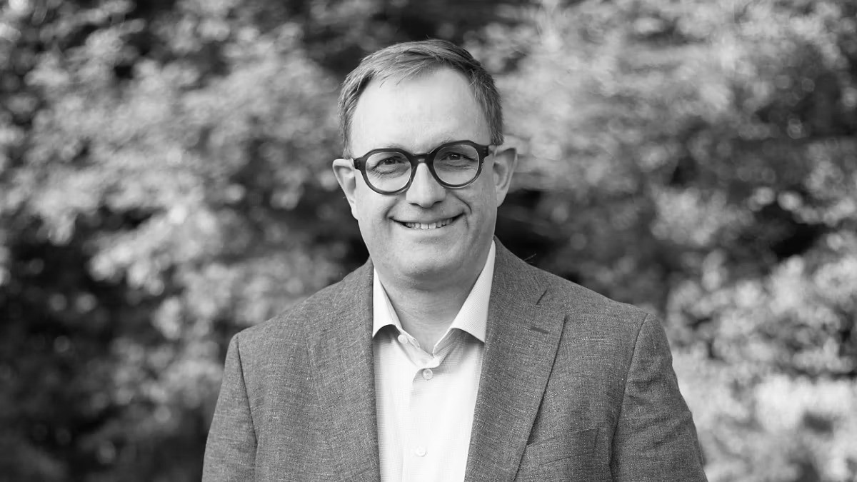 Homme souriant portant des lunettes et un veston, photographié en noir et blanc devant un fond d’arbres flous.