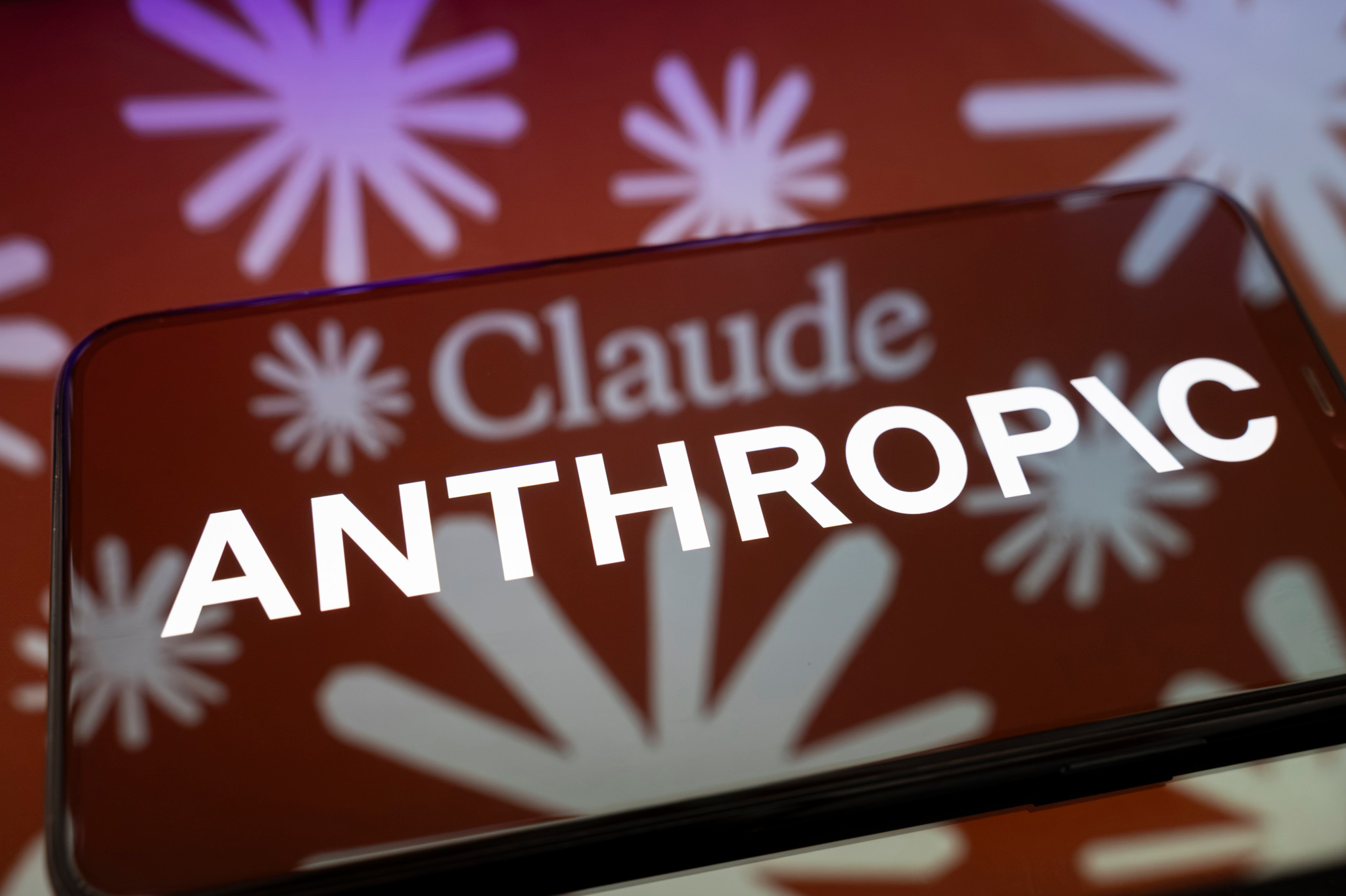 Logo Anthropic affiché sur un smartphone posé sur une surface réfléchissante, avec une illustration aux couleurs de l’IA Claude en arrière-plan.