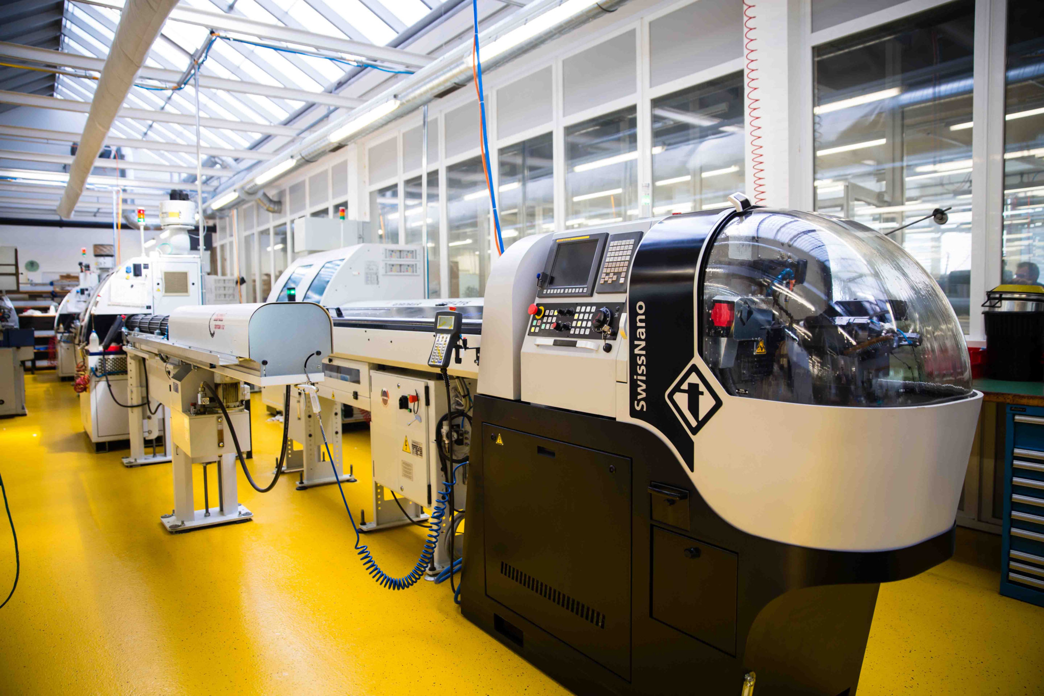 Atelier industriel avec machines-outils de précision SwissNano alignées sur sol jaune, dans une usine lumineuse à verrière.