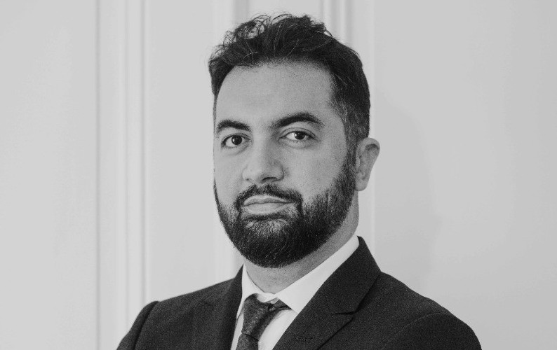 Portrait en noir et blanc d’un homme en costume et cravate, barbe soignée, regard sérieux vers l’objectif.