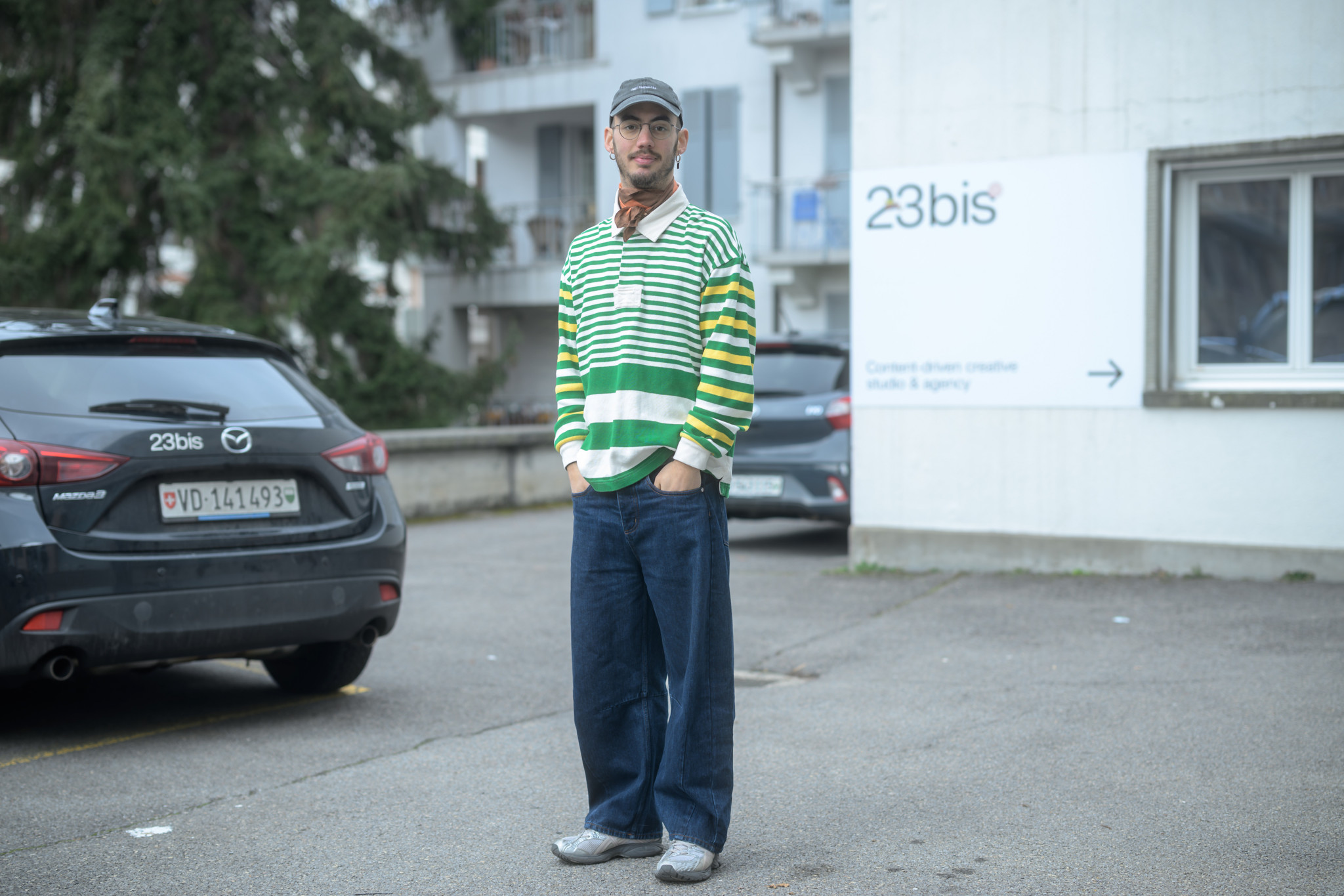 Gabriel Saurer debout devant le studio 23bis à Lausanne, portant un pull rayé vert et jaune, avec une voiture marquée du logo 23bis à gauche.