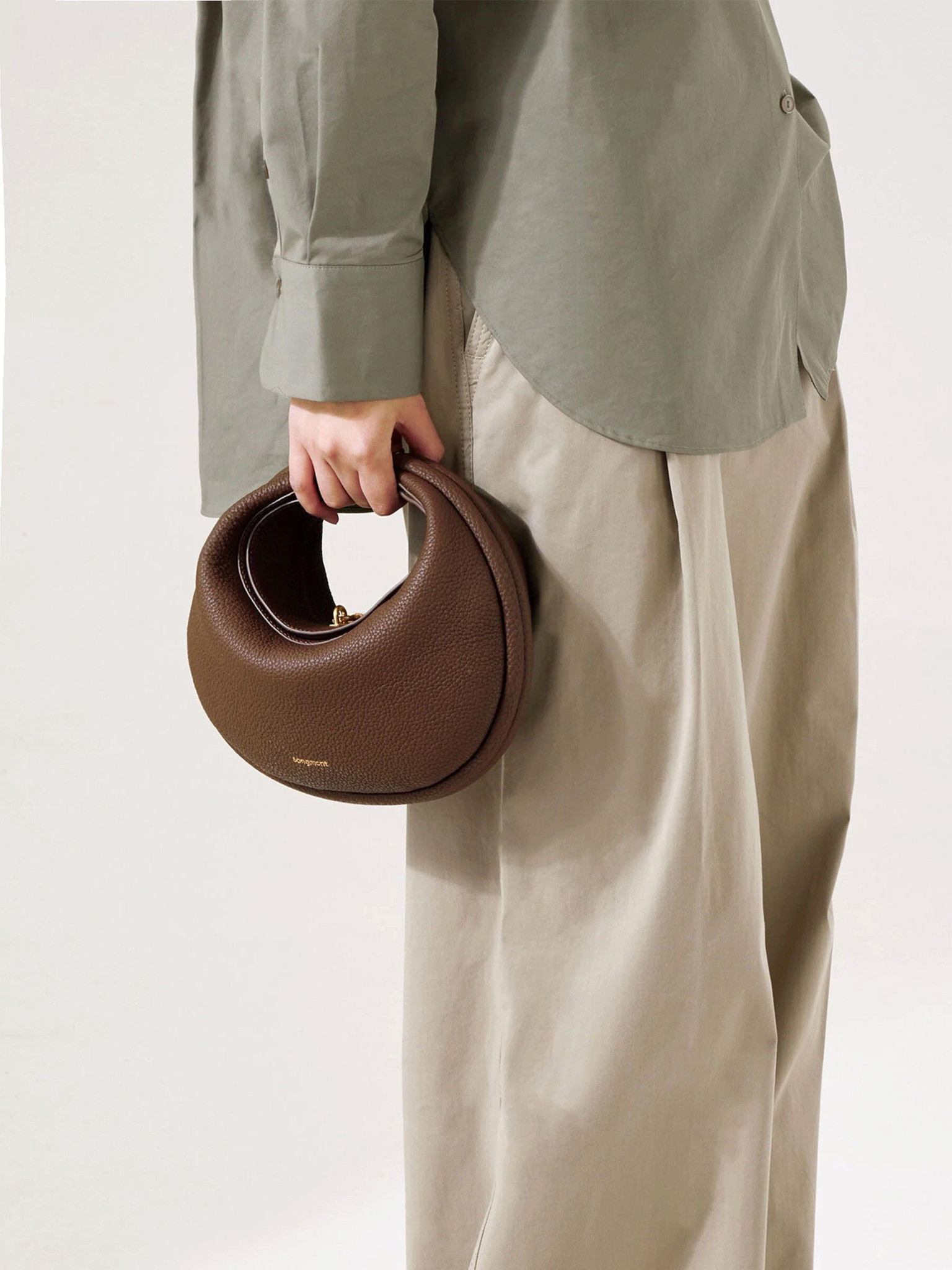 Femme tenant un sac à main en cuir marron de forme arrondie, portant une chemise verte et un pantalon beige ample.