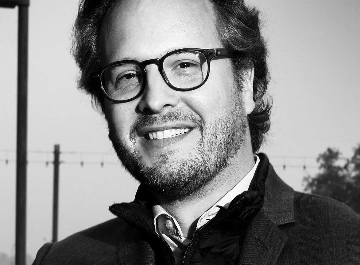 Portrait en noir et blanc d’un homme souriant avec des lunettes et une barbe courte, portant une veste.