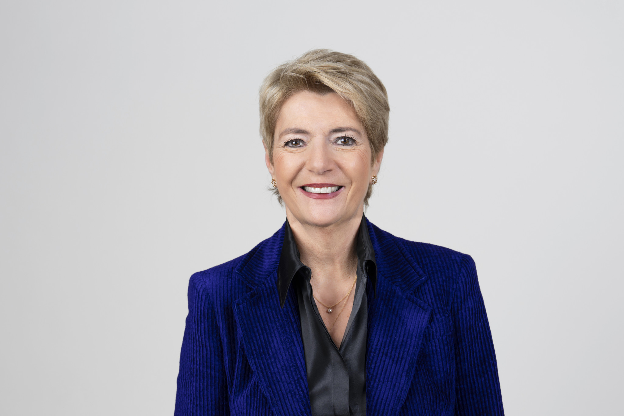 Femme aux cheveux courts blonds souriante, portant une veste bleue et une chemise noire, sur fond gris clair.