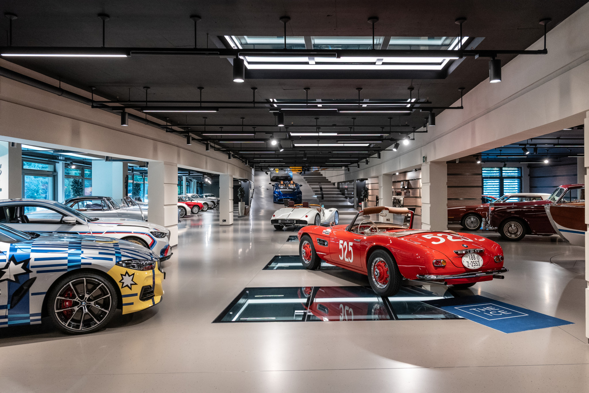 Salle d’exposition lumineuse avec plusieurs voitures de collection, dont une BMW rouge numéro 525 et une voiture aux couleurs vives artistiques.