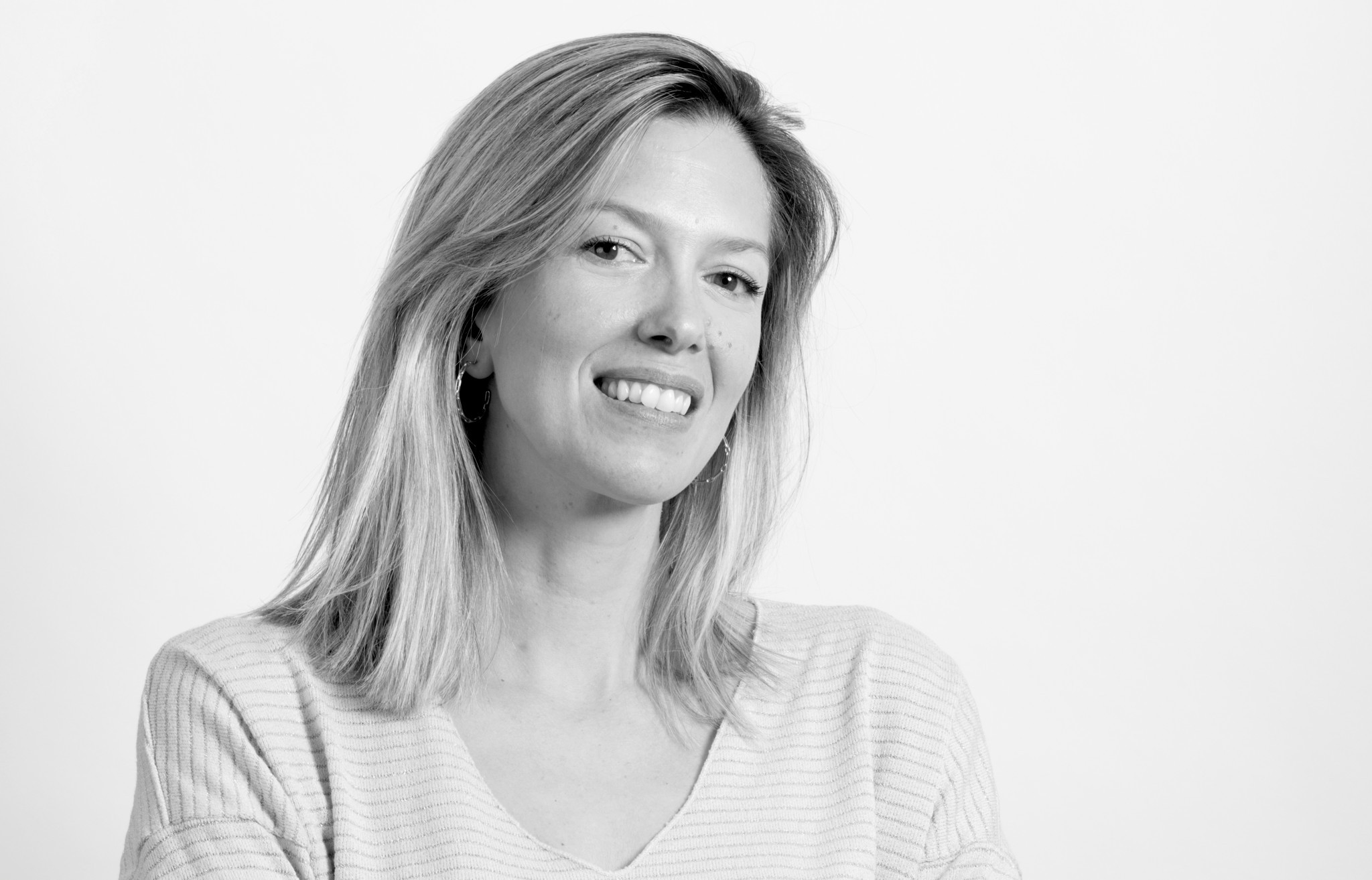 Portrait en noir et blanc d’une femme souriante aux cheveux blonds mi-longs, portant un haut rayé, sur fond clair.