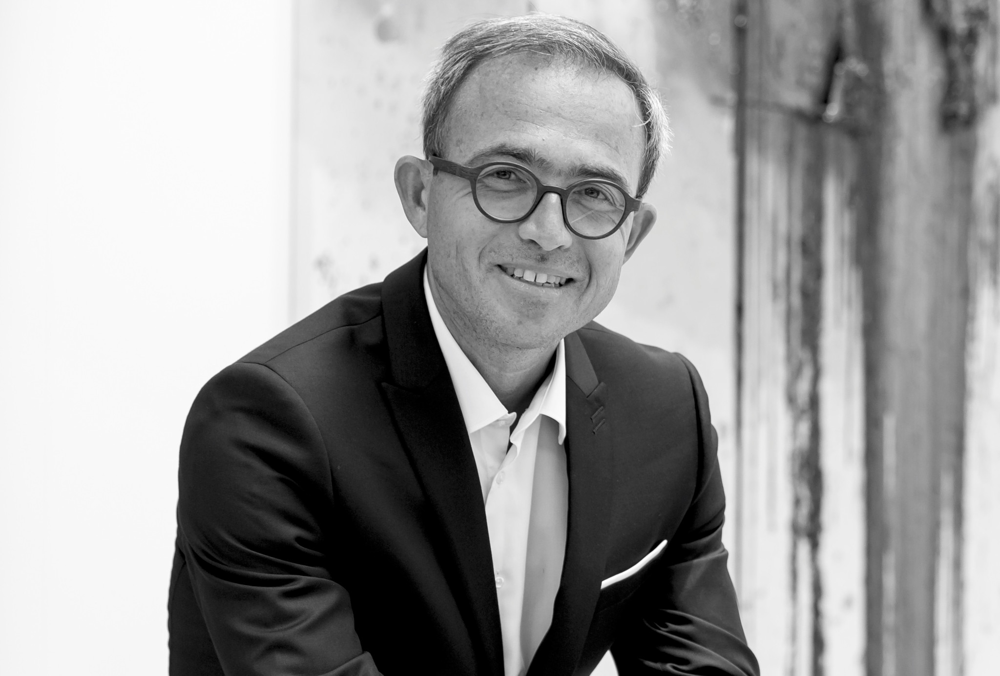 Portrait en noir et blanc d’un homme souriant en veste sombre et lunettes rondes, posant devant un fond texturé.