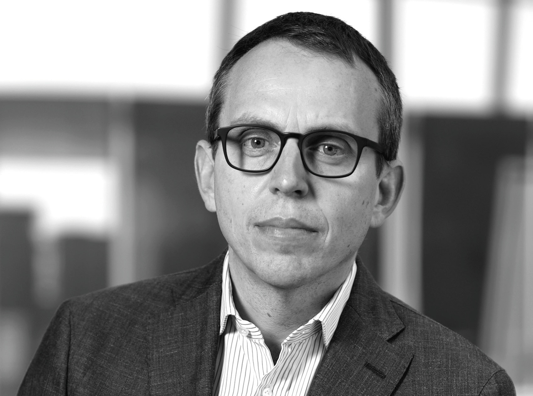 Portrait en noir et blanc d’un homme portant des lunettes et un veston, chemise à rayures, expression sérieuse.