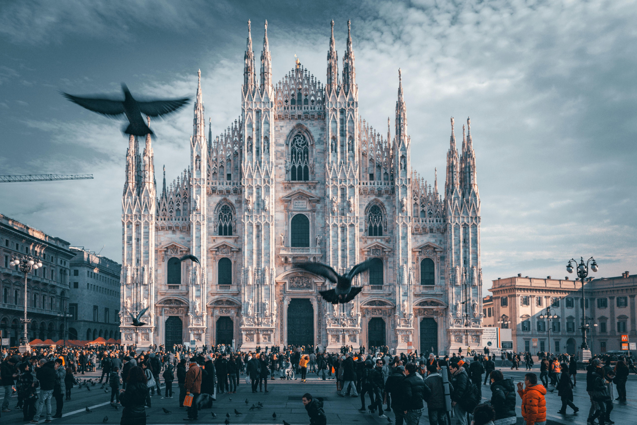 La cathédrale du Dôme de Milan avec des pigeons en vol, une foule animée sur la place et un ciel nuageux en arrière-plan.