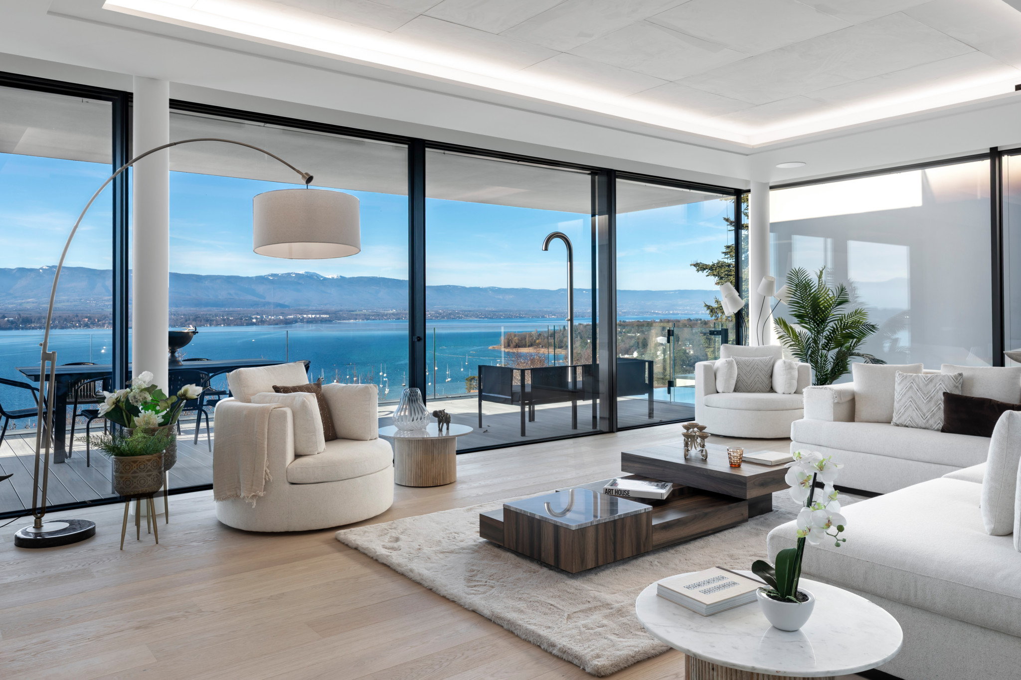 Salon luxueux aux tons blancs avec vue panoramique sur le lac Léman et les Alpes, baies vitrées, mobilier moderne et terrasse ensoleillée.