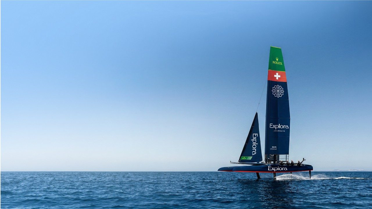 Catamaran de course Explora en pleine mer, voile verte ornée du logo Rolex et du drapeau suisse, naviguant sur foils par ciel bleu clair.