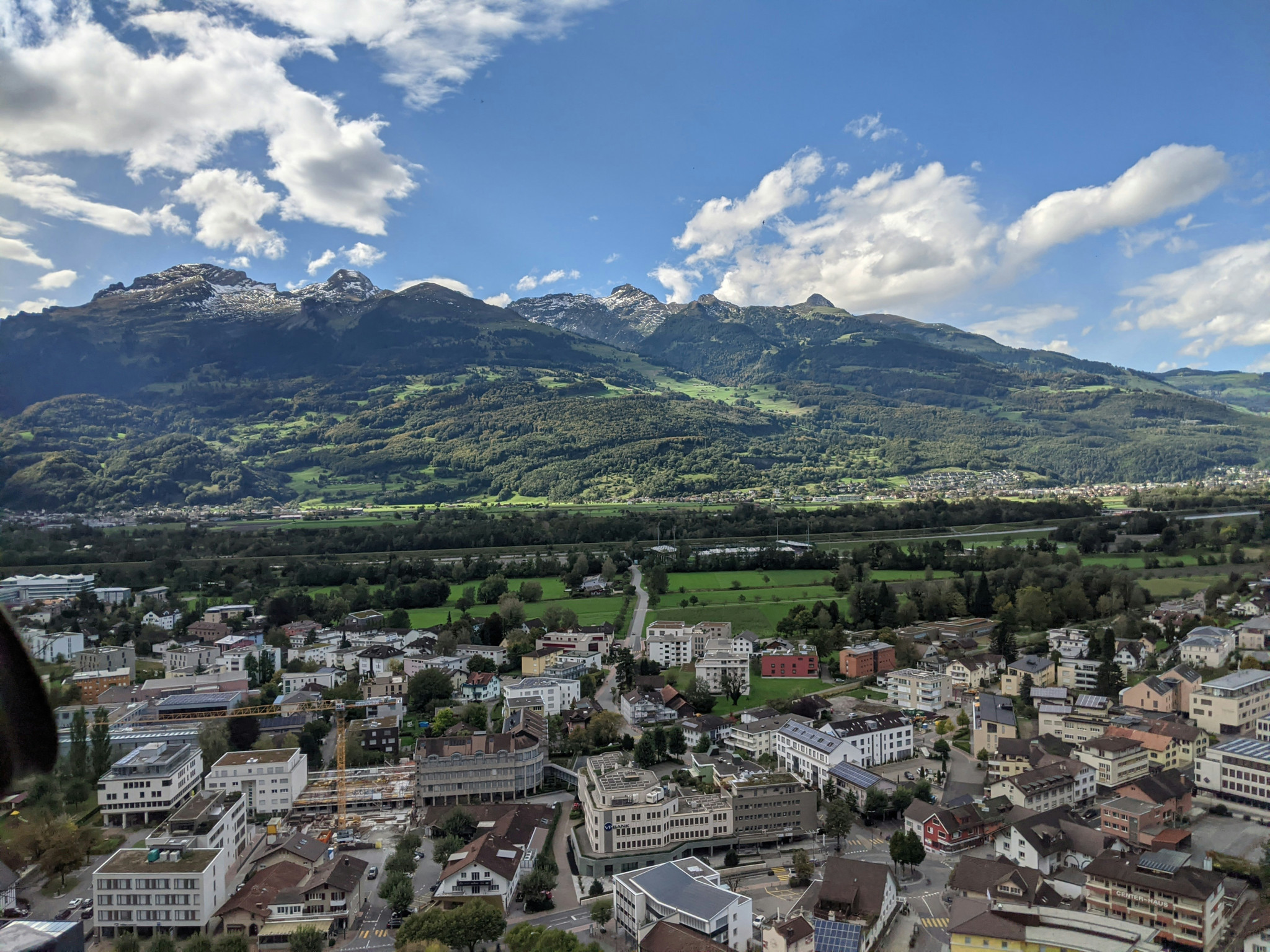 Vue aérienne d’une ville alpine avec bâtiments, vallée verdoyante et montagnes enneigées sous un ciel nuageux.