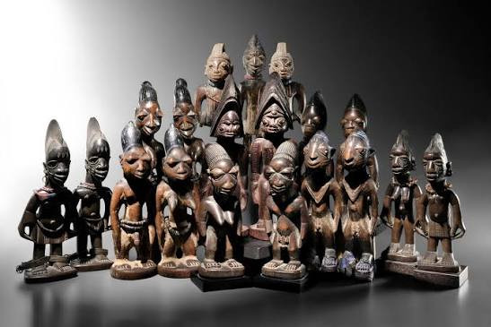Collection de statues africaines en bois sculpté, figurines Ibeji yoruba aux coiffures pointues, disposées en groupe sur fond gris.