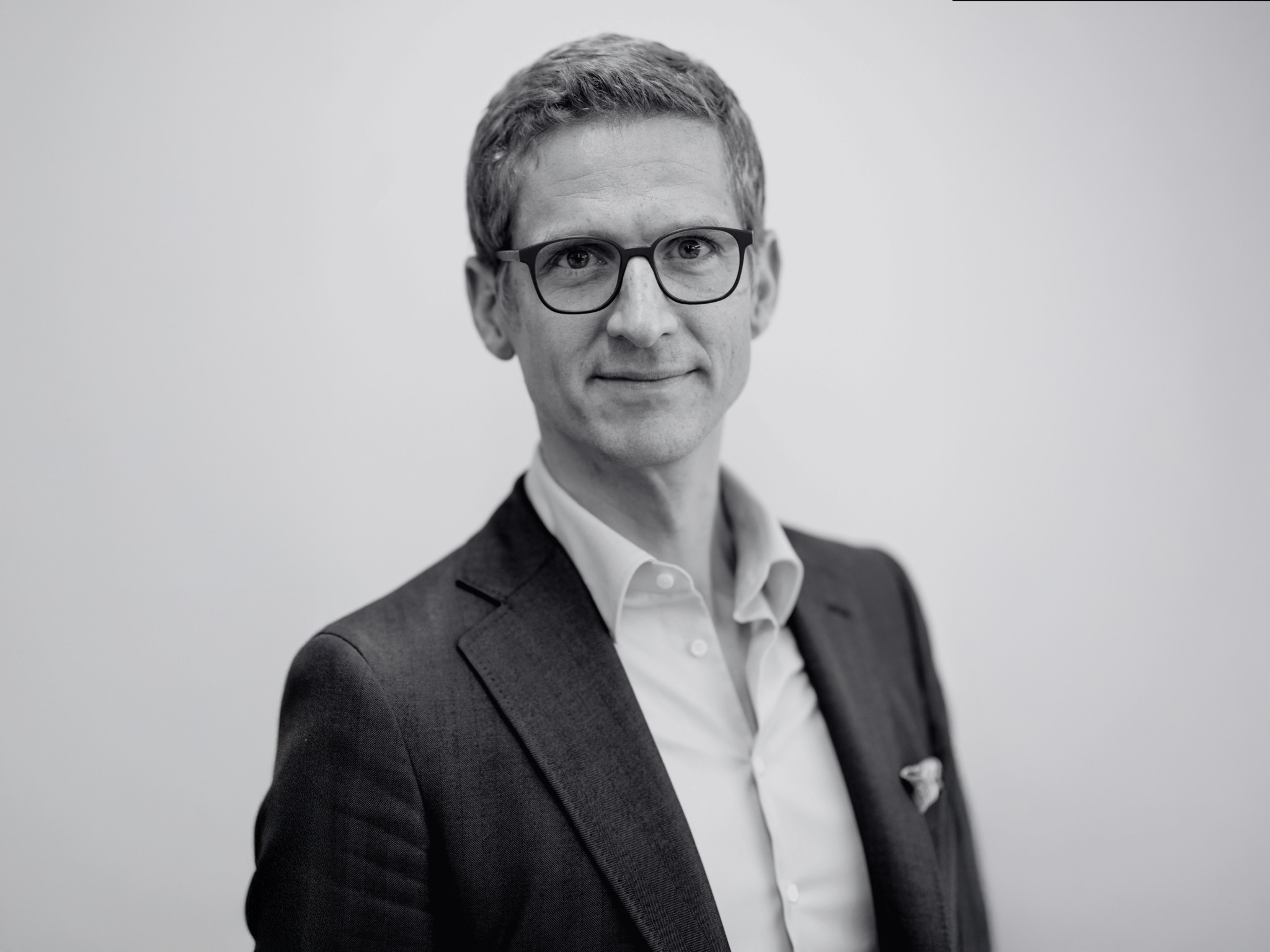 Portrait en noir et blanc d’un homme portant des lunettes et un costume sombre, chemise blanche, sur fond neutre.