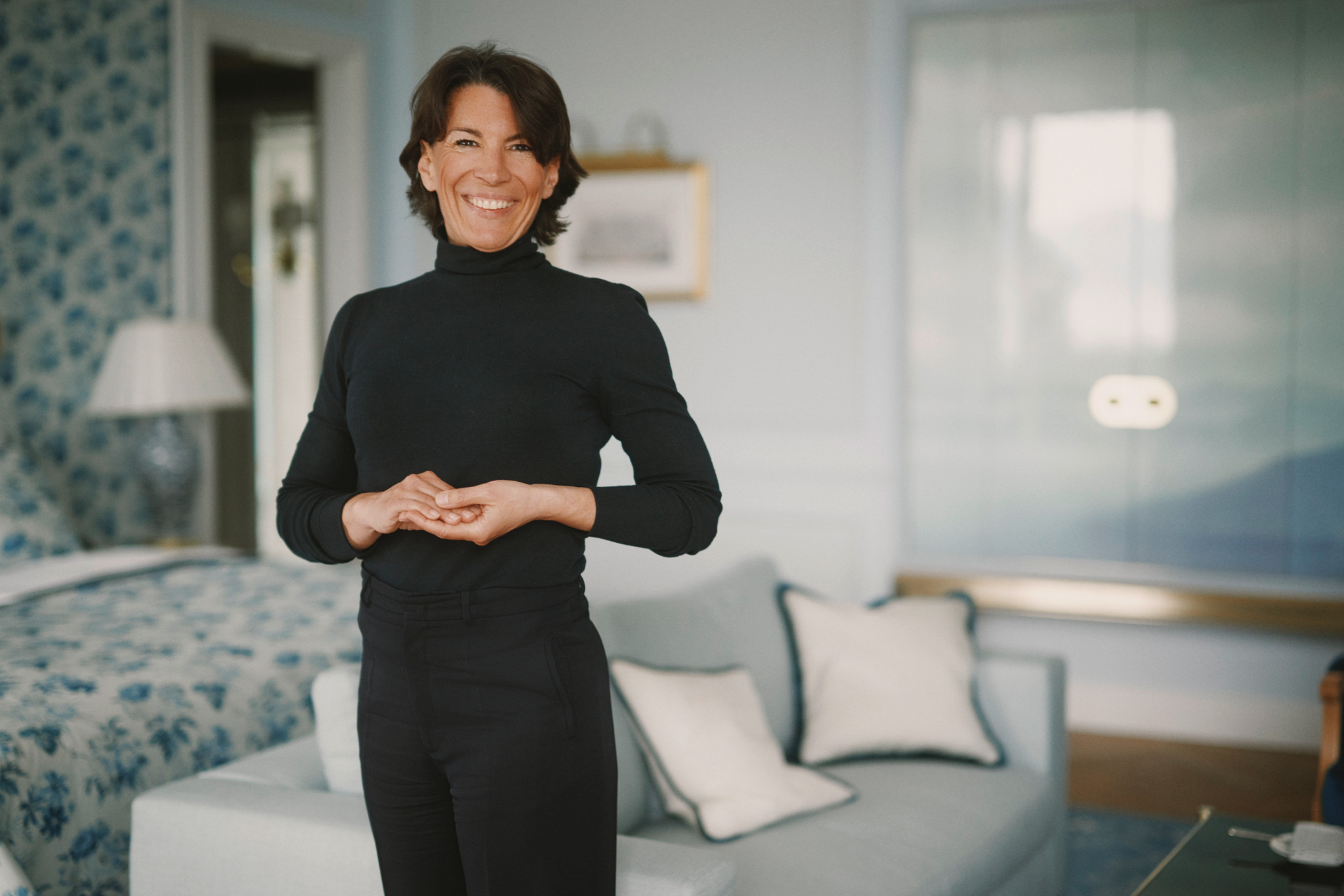 Portrait de Nathalie Seiler souriante, vêtue d’une tenue noire, dans un salon élégant aux tons bleus et blancs.