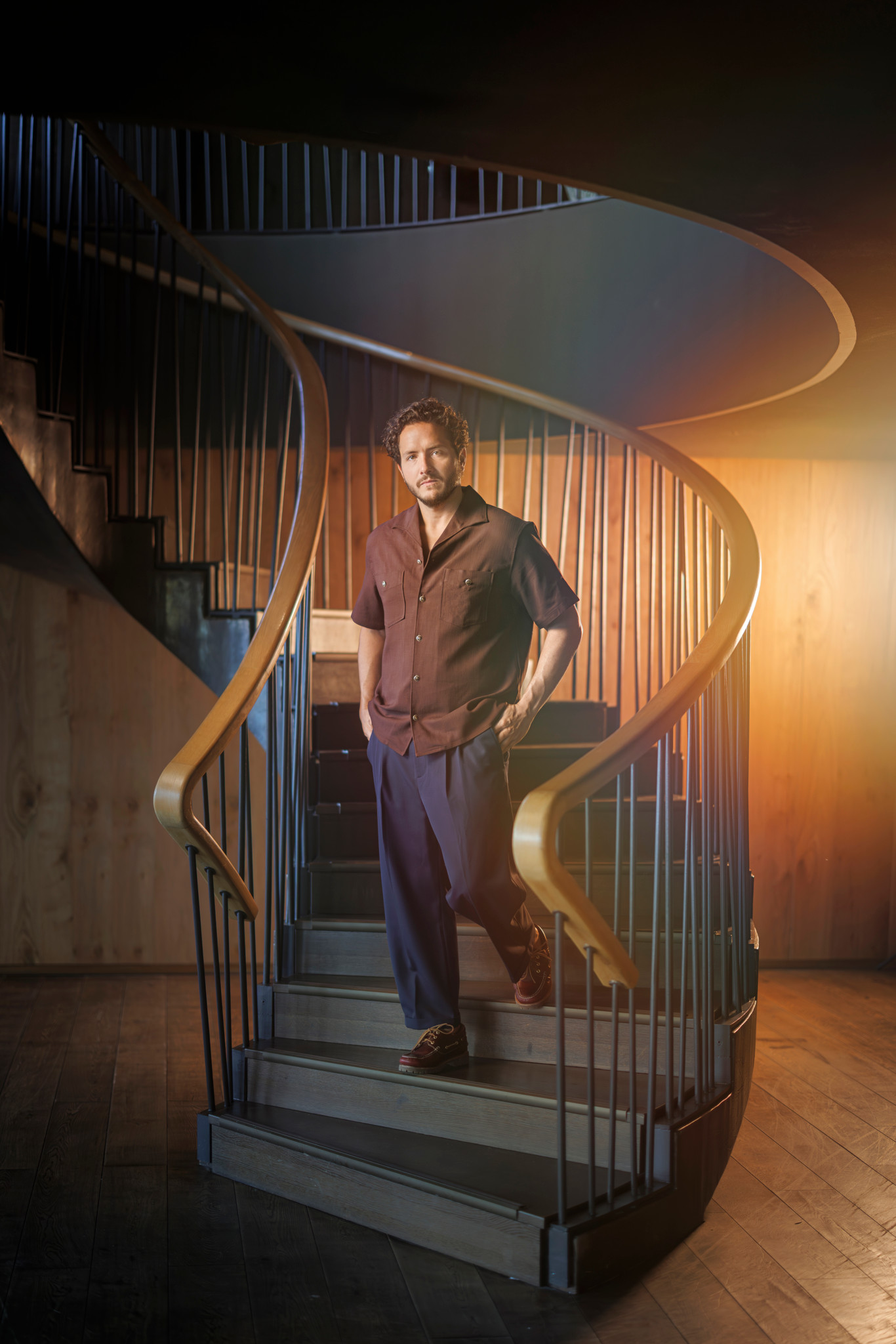 Alexandre Kominek posant sur un élégant escalier en colimaçon au design moderne, vêtu d’une chemise bordeaux et d’un pantalon bleu marine.