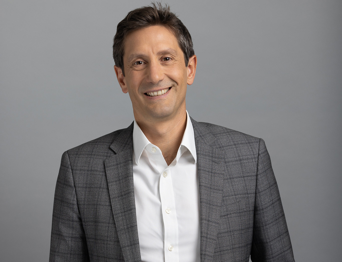 Homme souriant en costume gris avec une chemise blanche sur fond gris uni.