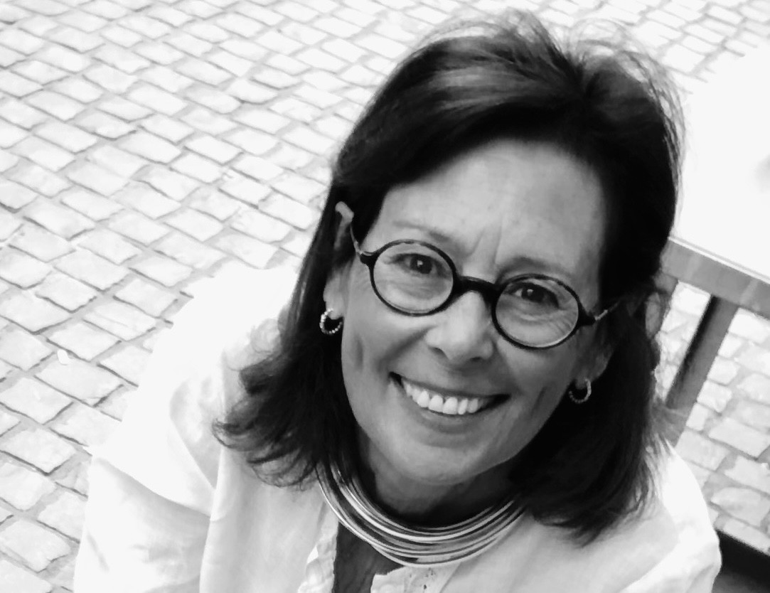 Femme souriante portant des lunettes rondes et une veste blanche, photographiée en noir et blanc sur fond de pavés.