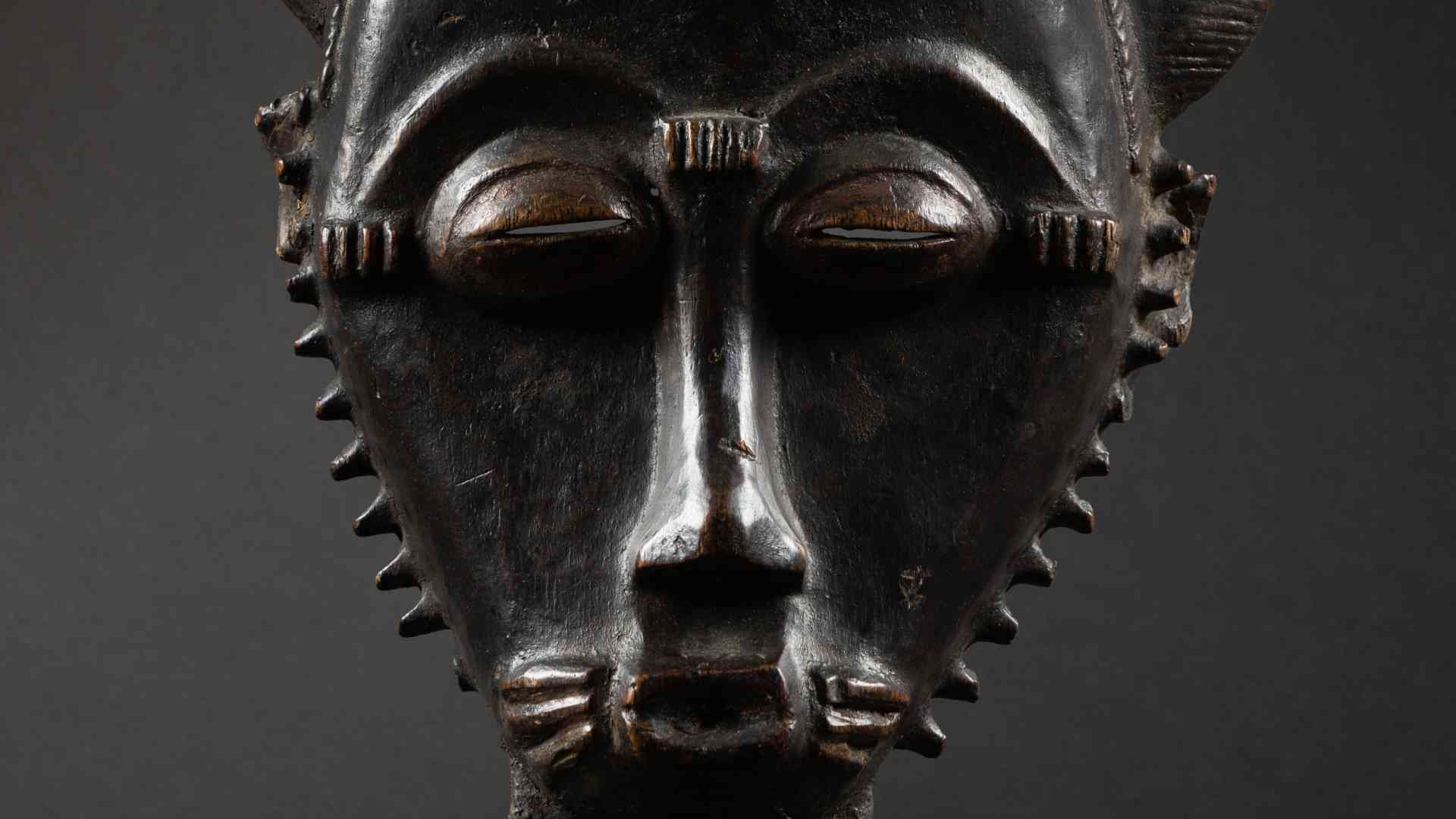 Masque africain traditionnel en bois sombre sculpté, aux traits fins et détaillés, yeux mi-clos et motifs géométriques, sur fond gris.