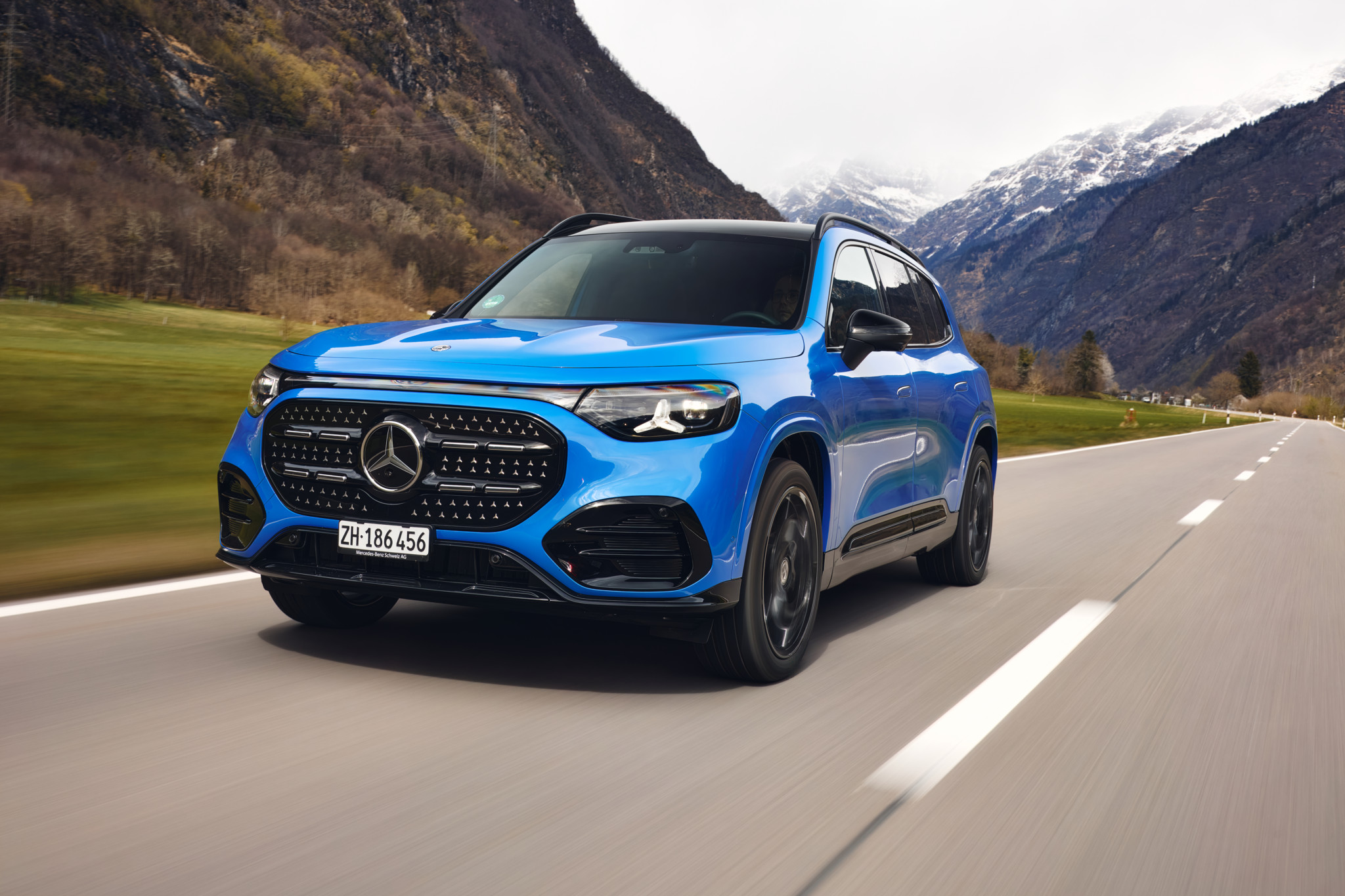 SUV Mercedes-Benz bleu en mouvement sur une route alpine, avec des montagnes enneigées en arrière-plan, plaque ZH 186 456.