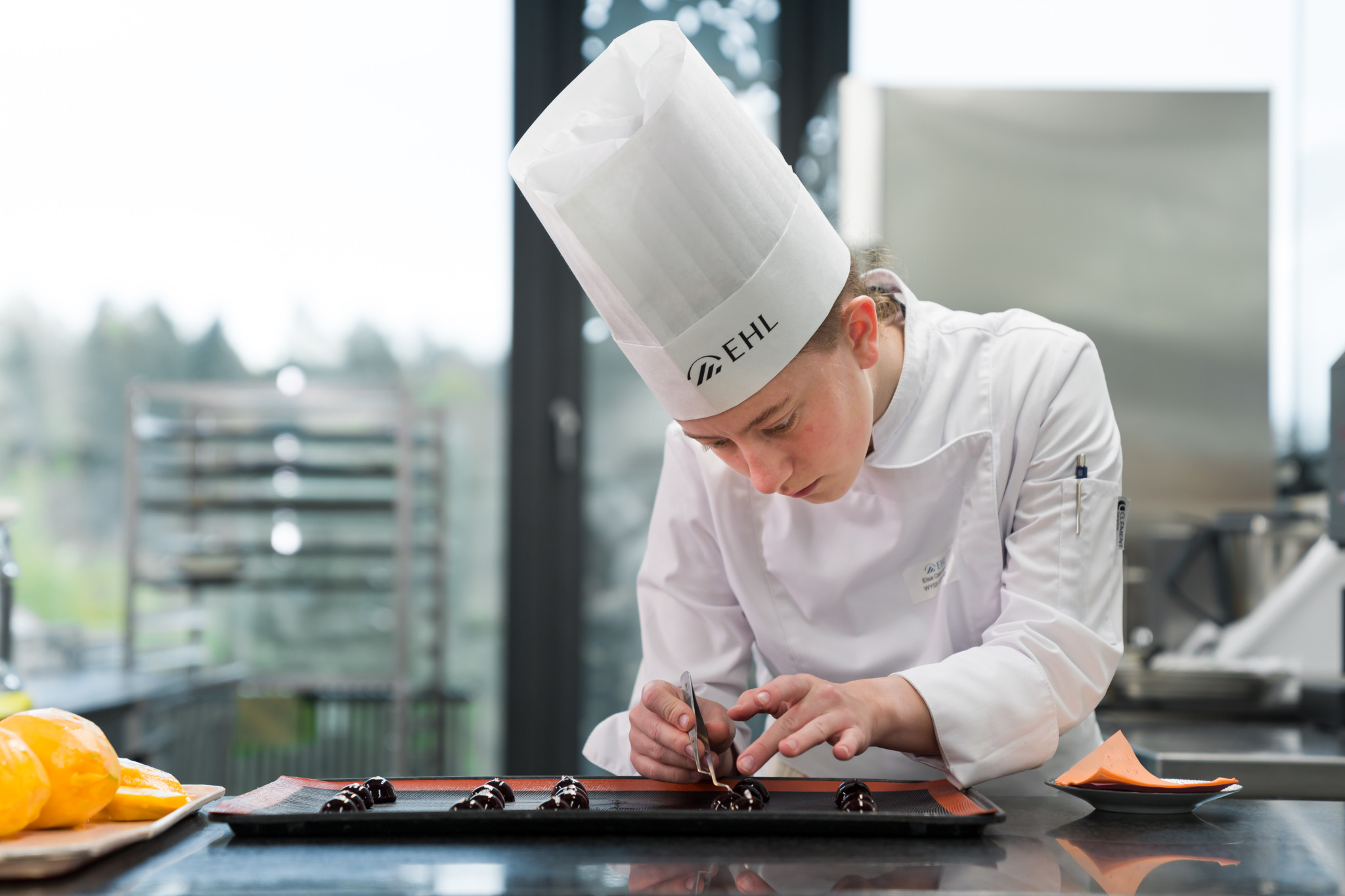 Étudiant·e de l’EHL en toque blanche décorant avec soin des chocolats sur un plateau dans une cuisine professionnelle.